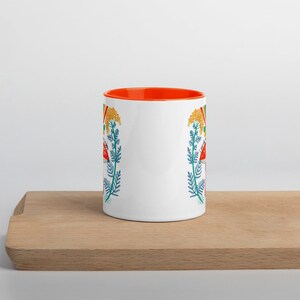 May include: White ceramic mug with an orange interior, featuring a colorful floral design with red mushrooms, yellow flowers, and blue foliage. The mug is displayed on a wooden cutting board.