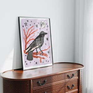 May include: A framed art print featuring a black crow perched on a red branch against a light purple background. The illustration includes a white moon and several black star shapes scattered throughout the composition, all enclosed in a thin black frame.