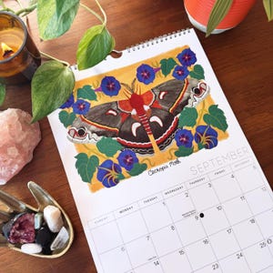 May include: A spiral-bound calendar featuring a colorful illustration of a Cecropia Moth surrounded by blue flowers and green leaves. The calendar is open to September, with the month's name and days of the week visible.