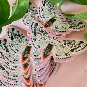 May include: A stack of butterfly stickers with a pink border, white wings, and black and pink details. The stickers have the text "@DREAM FOLK STUDIO". Green leaves are in the background.