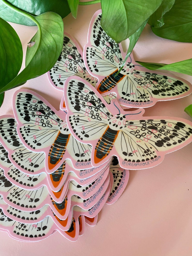 May include: A stack of butterfly stickers with a pink border, white wings, and black and pink details. The stickers have the text "@DREAM FOLK STUDIO". Green leaves are in the background.