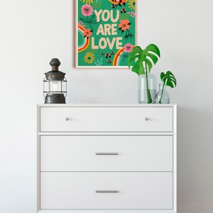 Pu&ograve; includere: Una cassettiera bianca con tre cassetti e una stampa incorniciata che dice "You are Love" con un arcobaleno e un design floreale su sfondo verde.