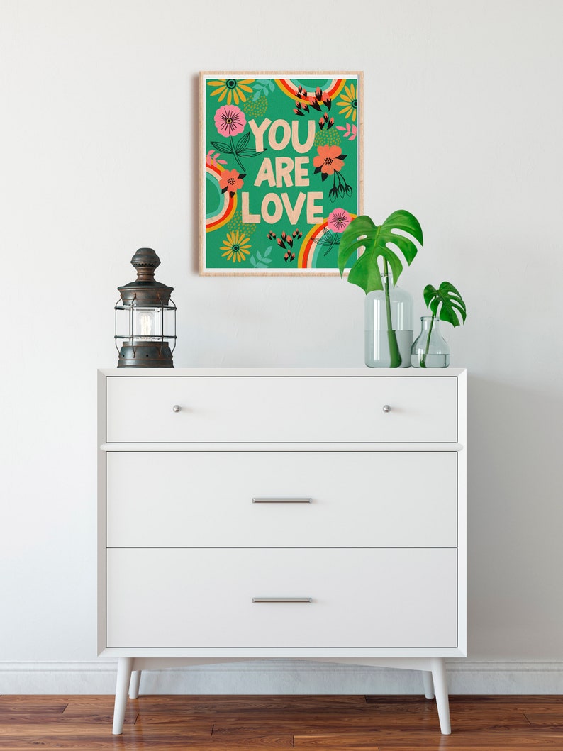 Pu&ograve; includere: Una cassettiera bianca con tre cassetti e una stampa incorniciata che dice "You are Love" con un arcobaleno e un design floreale su sfondo verde.
