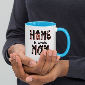 K&ouml;nnte beinhalten: Eine wei&szlig;e Keramiktasse mit blauem Rand und Henkel. Die Tasse hat ein schwarzes und pinkfarbenes Design mit dem Schriftzug "Home is where Mom is".