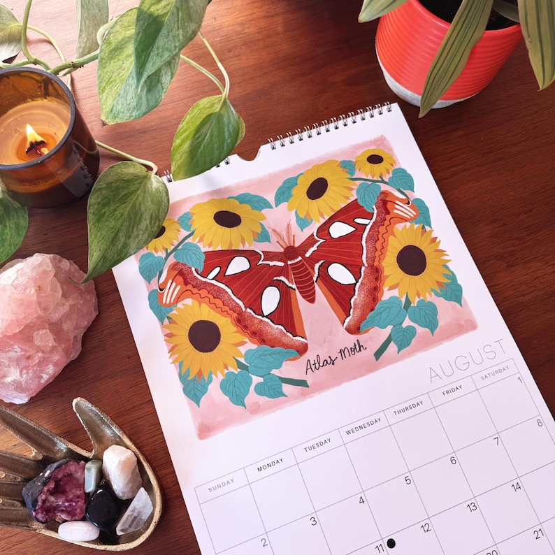May include: A spiral-bound calendar open to August, featuring an illustration of an Atlas Moth surrounded by sunflowers. The moth is red and orange with white markings. The calendar is on a wooden surface with plants and a candle.