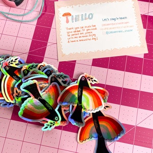 May include: A stack of iridescent rainbow bird stickers with black outlines and a small green leaf on each bird. The stickers are on a pink cutting mat with a white grid pattern. The text on the card reads "HELLO" and "Let's stay in touch: DREAM FOLKSTUDIO.com HELLO@DREAM FOLKSTUDIO.com @DREAM FOLK_STUDIO"