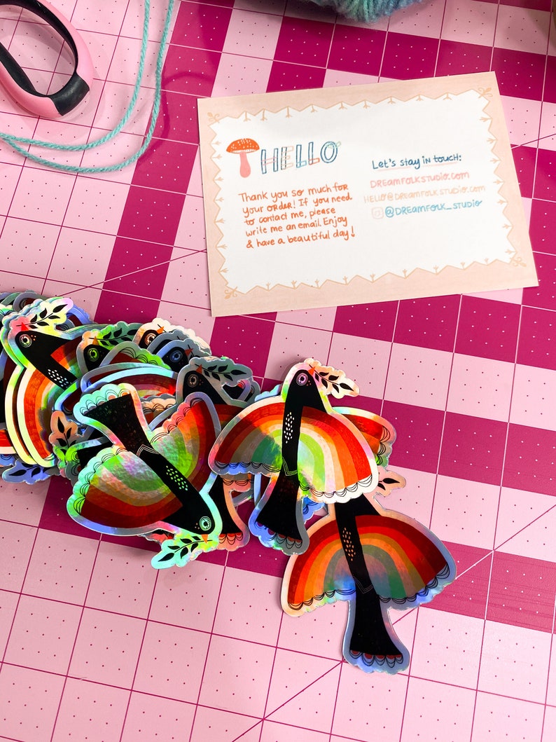 May include: A stack of iridescent rainbow bird stickers with black outlines and a small green leaf on each bird. The stickers are on a pink cutting mat with a white grid pattern. The text on the card reads "HELLO" and "Let's stay in touch: DREAM FOLKSTUDIO.com HELLO@DREAM FOLKSTUDIO.com @DREAM FOLK_STUDIO"