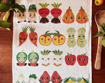 May include: A rectangular off-white kitchen towel featuring a repeating pattern of anthropomorphic fruits and vegetables with faces. The top and bottom borders display a colorful striped design. The text Dream Folk Studio is printed at the bottom.