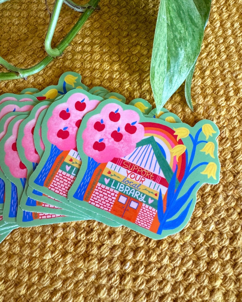 May include: A stack of green and pink stickers with a colorful illustration of a house with a rainbow, flowers, and the text "Support your local library".