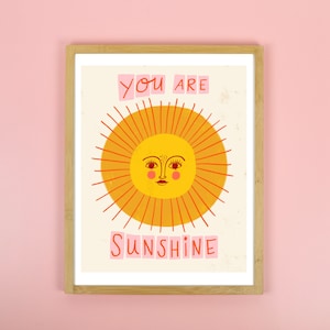 K&ouml;nnte beinhalten: Ein gerahmter Druck mit einer gelben Sonne mit einem l&auml;chelnden Gesicht und dem Text "You are Sunshine" in rosa Buchstaben.