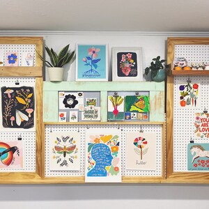 May include: A wall display of framed artwork featuring colourful illustrations of flowers, insects, and other nature-inspired designs. The artwork is displayed on pegboards and shelves, creating a vibrant and eclectic gallery wall.