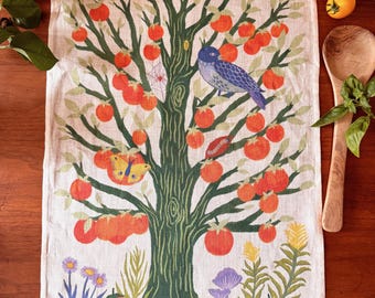 May include: A rectangular kitchen towel featuring a central dark green tree with orange tomato-like fruits, a blue bird, a yellow butterfly, and a spider web. The base shows purple flowers, green plants, and a snail on a yellow horizontal band.