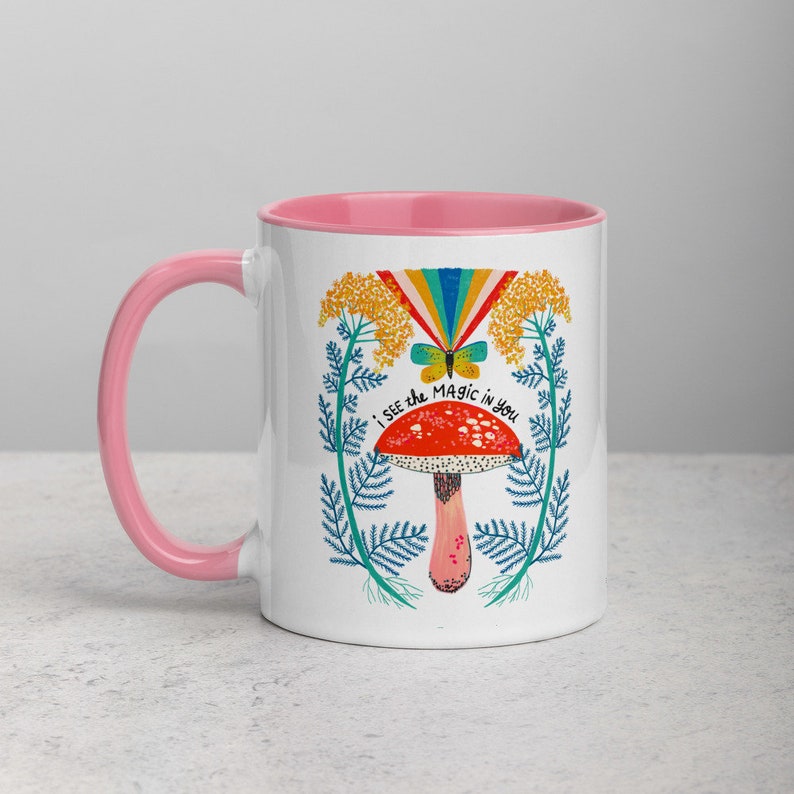 May include: White ceramic mug with a pink interior and handle. The mug features a colorful illustration of a mushroom, butterfly, and rainbow with the text "I SEE THE MAGIC IN YOU". The design is surrounded by blue and yellow floral elements.