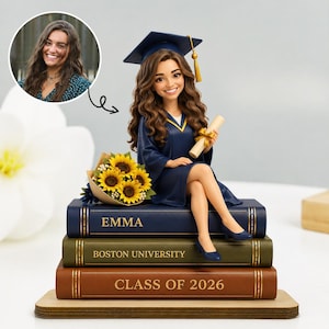 May include: A custom figurine of a graduate with long brown hair wearing a navy blue cap and gown, holding a diploma. The figure sits on a stack of three books labeled EMMA, BOSTON UNIVERSITY, and CLASS OF 2026, next to a bouquet of yellow sunflowers.