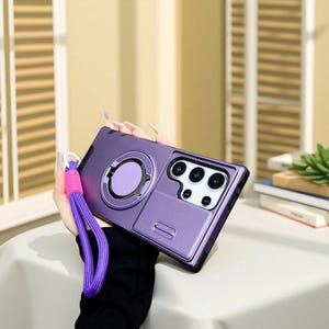 May include: A purple phone case with a ring holder and a camera protector. The case has a matching purple ring holder and a sliding camera cover. A purple and pink wrist strap is attached to the case.