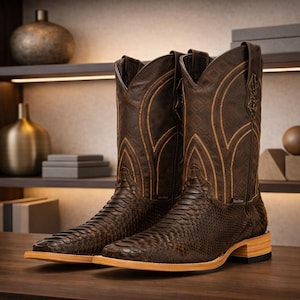 May include: A pair of brown leather cowboy boots with a snake-skin pattern. The boots have a square toe and a light brown sole. The shaft of the boots features decorative stitching in a lighter brown color. The boots are displayed on a dark wooden surface.