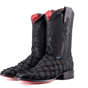 May include: A pair of black cowboy boots with a textured, scale-like pattern. The boots have a red interior and soles. The design includes decorative stitching and a square toe. The boots are set against a white background.