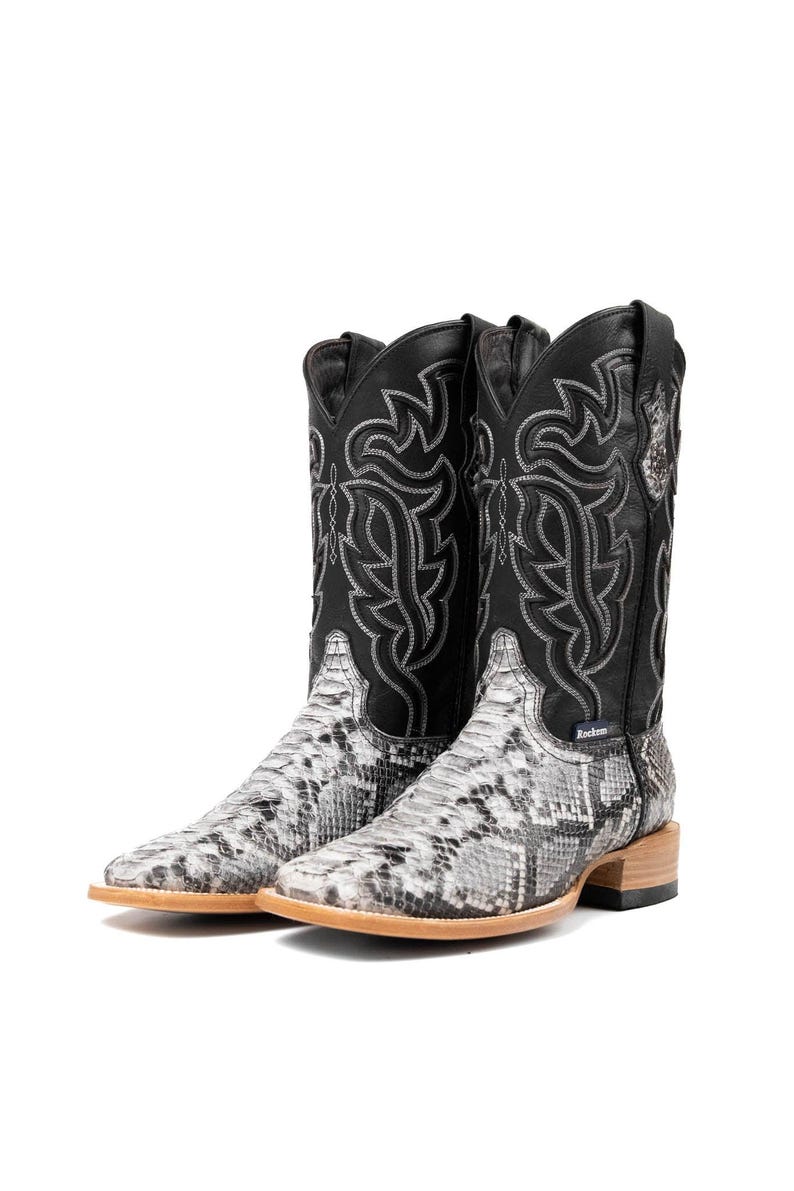 May include: A pair of cowboy boots with a snakeskin pattern on the foot and a black leather upper. The black upper features decorative stitching in a silver color. The boots have a square toe and a light brown sole.
