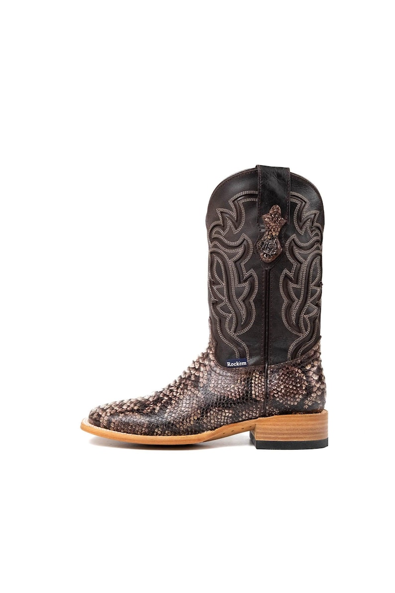 May include: A brown and tan cowboy boot with a snakeskin pattern on the lower portion and a decorative stitched design on the upper shaft. The boot has a square toe and a wooden heel. The brand name "Rockem" is visible.