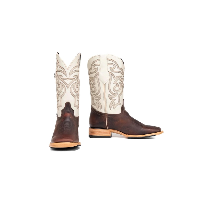 May include: A pair of brown and white cowboy boots. The boots have a square toe and a light-coloured sole. The shaft is white with decorative stitching, and the foot is a dark brown leather.