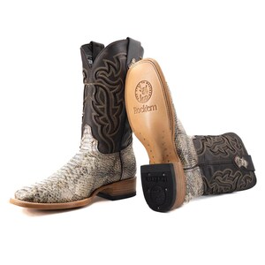 May include: A pair of cowboy boots with a snakeskin pattern on the foot and a dark brown leather upper. The boots have a square toe and decorative stitching. The soles are visible, with the brand name "Rock'em" printed on them.