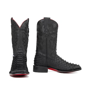 Puede incluir: Botas de vaquero negras con un patrón texturizado y escamoso en el pie y la parte inferior de la pierna. Las botas tienen suela y forro rojos, con costuras decorativas en la caña superior. Fondo blanco.