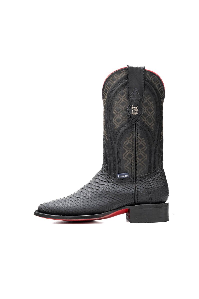 May include: Black cowboy boot with a textured snakeskin pattern on the foot and a decorative stitched shaft. The boot has a red sole and a small metal emblem near the top. The brand name "Rockem" is visible.