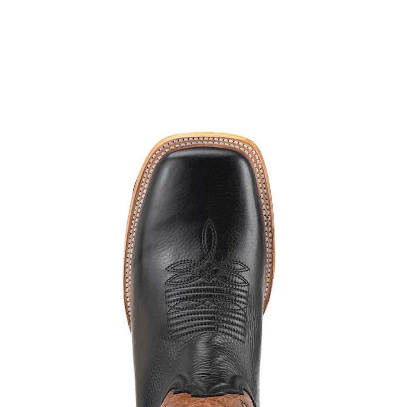 May include: Overhead shot of a black leather cowboy boot with a square toe. The boot has decorative stitching on the vamp and a light brown sole. The upper part of the boot is black, and the sole is light brown.
