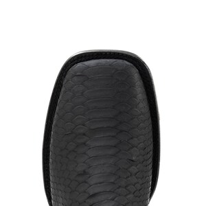 May include: Close-up of a black leather boot with a square toe. The boot features a textured, scale-like pattern across the surface. The top edge of the boot is smooth and black.