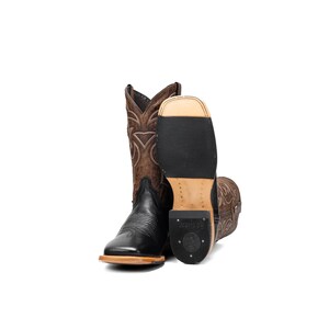May include: A pair of cowboy boots, one upright and the other lying on its side. The boots are primarily brown and black, with intricate stitching and a square toe design. The soles are black and the heels are a light brown colour.