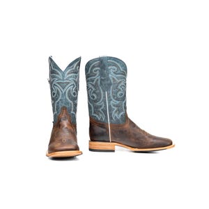 May include: A pair of brown leather cowboy boots with a square toe. The boots feature a blue and white embroidered design on the shaft and a brown leather foot. The boots are set against a white background.