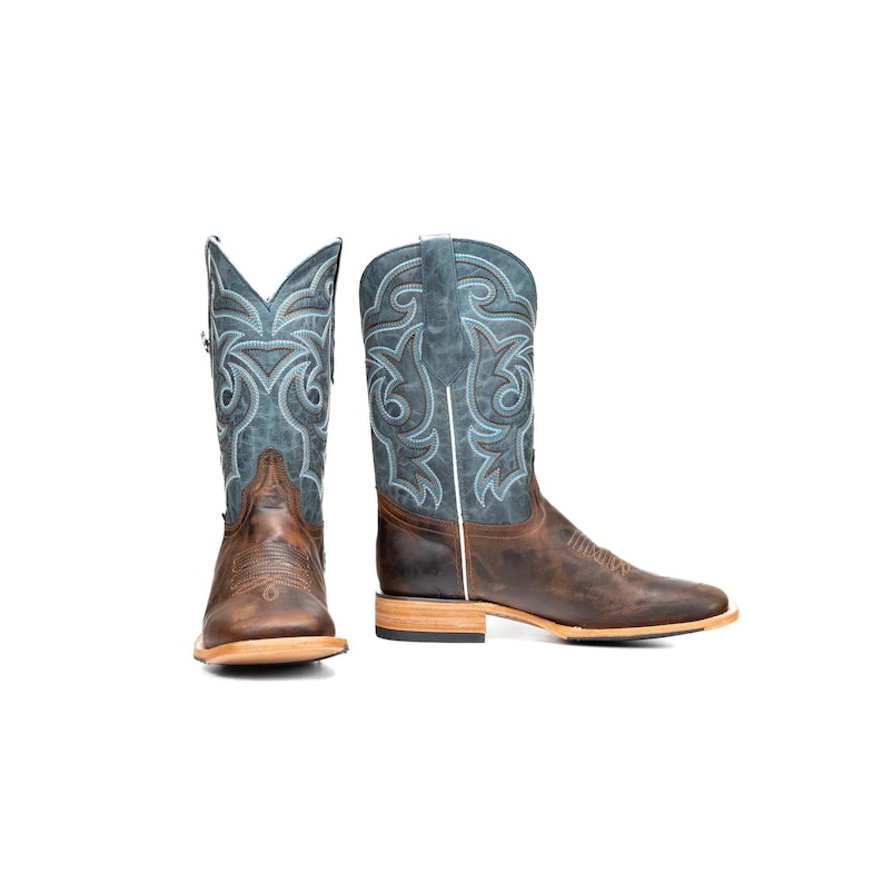 May include: A pair of brown leather cowboy boots with a square toe. The boots feature a blue and white embroidered design on the shaft and a brown leather foot. The boots are set against a white background.