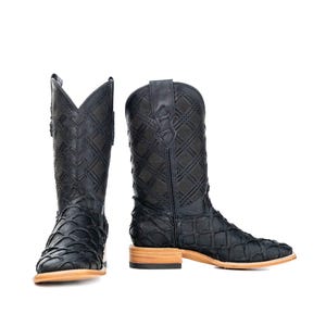Può includere: Stivali da cowboy in pelle nera con punta quadrata e motivo decorativo cucito. Gli stivali hanno un design strutturato e una suola marrone chiaro. Gli stivali sono esposti su sfondo bianco.