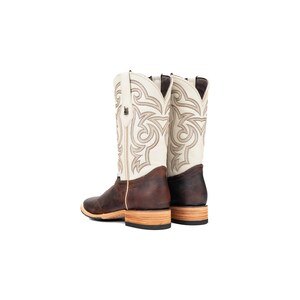 May include: A pair of cowboy boots. They have a white upper with detailed stitching, a brown leather foot, and a light-coloured wooden sole. The boots have a classic Western design.