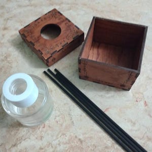 May include: A wooden tissue box with a decorative floral design, a glass diffuser bottle with a white top, and black diffuser sticks. The box is open, revealing the interior. The items are arranged on a light-colored surface.