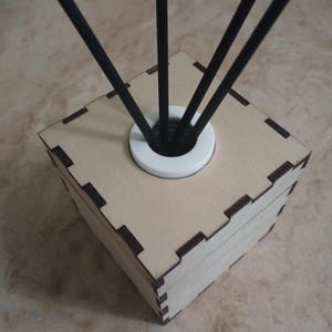 May include: A light-colored wooden cube-shaped diffuser with a white circular top. Black reeds extend upwards from the center. The cube has dark brown accents at the corners and edges. The background is a light brown surface.