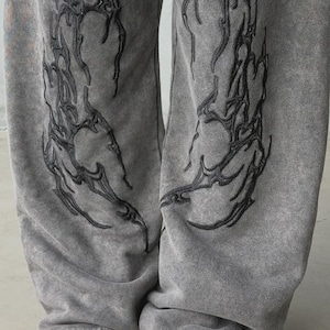 Puede incluir: Pantalones deportivos grises con bordados negros de estilo tribal en las piernas. Los pantalones tienen un aspecto descolorido y vintage y se combinan con zapatillas negras y blancas. El diseño presenta un patrón abstracto único.