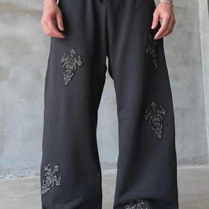 May include: Black sweatpants with a wide leg design. The pants feature decorative embroidered cross-like patterns in a lighter color. The waistband has a drawstring. The pants are made of a soft material.