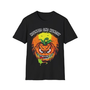 May include: Black t-shirt featuring a Halloween-themed graphic. The design includes a grinning jack-o'-lantern with sharp teeth, green slime, and the text "HAUNTED AND HUNGRY" in a stylized font. The pumpkin is set against an orange background.