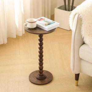 Puede incluir: Una mesa auxiliar de madera marrón oscuro con una parte superior y una base redondas, conectadas por una columna de cuentas. Una taza blanca con borde marrón y un libro titulado "Delicious Places" descansan sobre la mesa. La mesa está al lado de un sofá color crema.