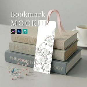 May include: A rectangular white bookmark featuring a delicate grey floral vine pattern and a light pink ribbon attached to the top. The bookmark is displayed standing upright against a stack of three hardcover books with visible text.