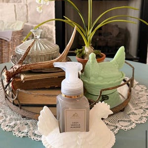 May include: A decorative display featuring a white ceramic hen soap holder with a gray soap dispenser labeled "Cozy Vanilla Almond." The arrangement includes a green hen-shaped dish, a tray with books, and a glass jar, creating a farmhouse aesthetic.