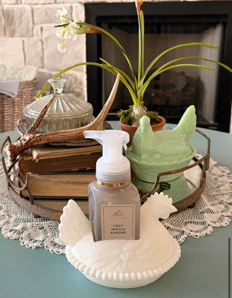 May include: A decorative display featuring a white ceramic hen soap holder with a gray soap dispenser labeled "Cozy Vanilla Almond." The arrangement includes a green hen-shaped dish, a tray with books, and a glass jar, creating a farmhouse aesthetic.