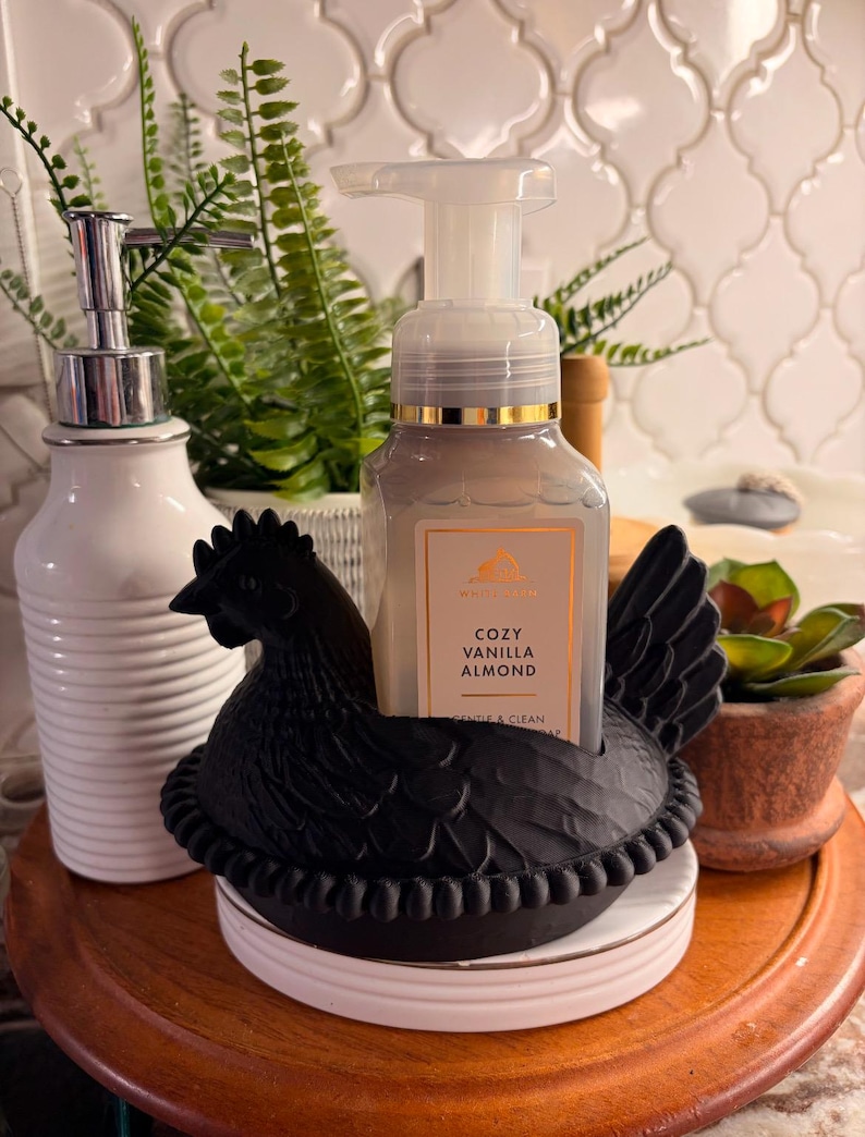 May include: A black hen-shaped soap holder with a bottle of Cozy Vanilla Almond soap. The soap dispenser is clear with a gold-colored rim. A white ceramic soap dispenser and potted plants are also visible.