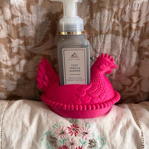May include: A gray hand soap bottle labeled "Cozy Vanilla Almond" sits in a bright pink chicken-shaped holder. The soap dispenser has a white pump. The holder is on a white surface with floral embroidery.