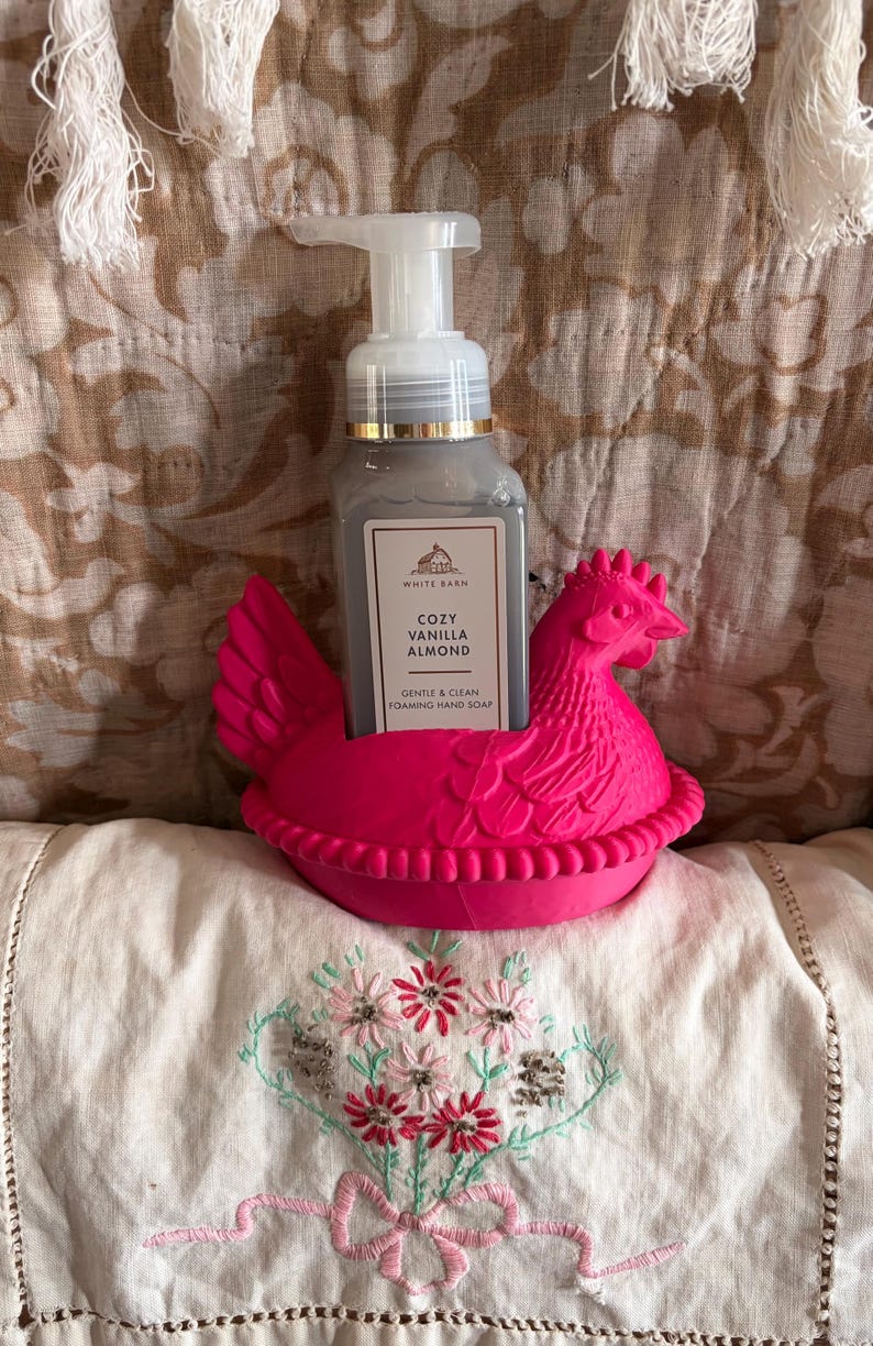 May include: A gray hand soap bottle labeled "Cozy Vanilla Almond" sits in a bright pink chicken-shaped holder. The soap dispenser has a white pump. The holder is on a white surface with floral embroidery.