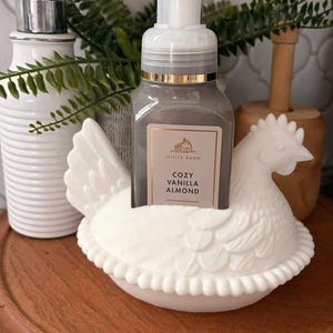 May include: A white ceramic chicken-shaped soap holder cradles a gray soap dispenser with a gold rim. The soap bottle has a label that reads "Cozy Vanilla Almond." A white, ribbed soap dispenser and greenery are in the background.