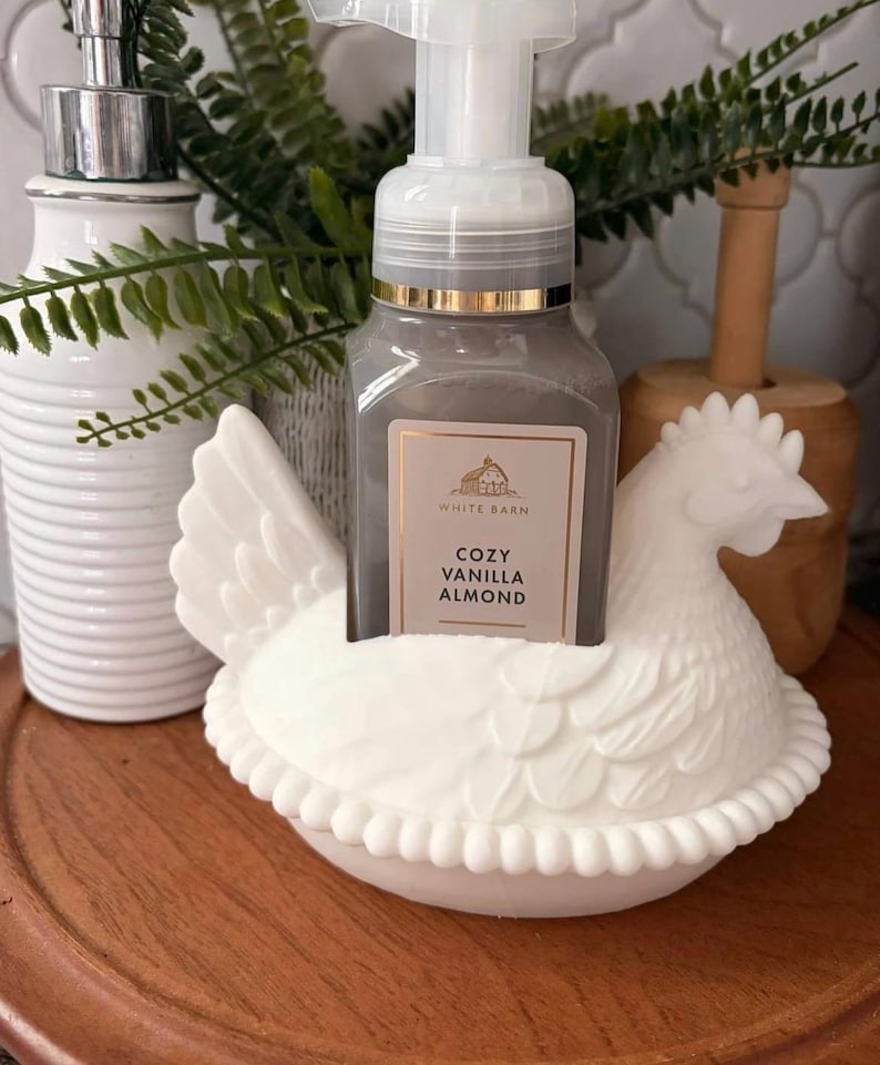May include: A white ceramic chicken-shaped soap holder cradles a gray soap dispenser with a gold rim. The soap bottle has a label that reads "Cozy Vanilla Almond." A white, ribbed soap dispenser and greenery are in the background.
