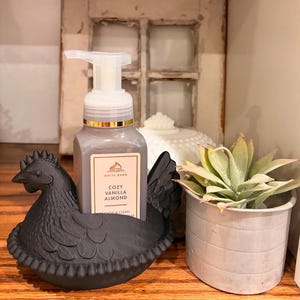 May include: A black hen-shaped soap dispenser holder cradles a bottle of Cozy Vanilla Almond soap. A small potted succulent and a decorative white object are also visible. The background includes a distressed window frame.