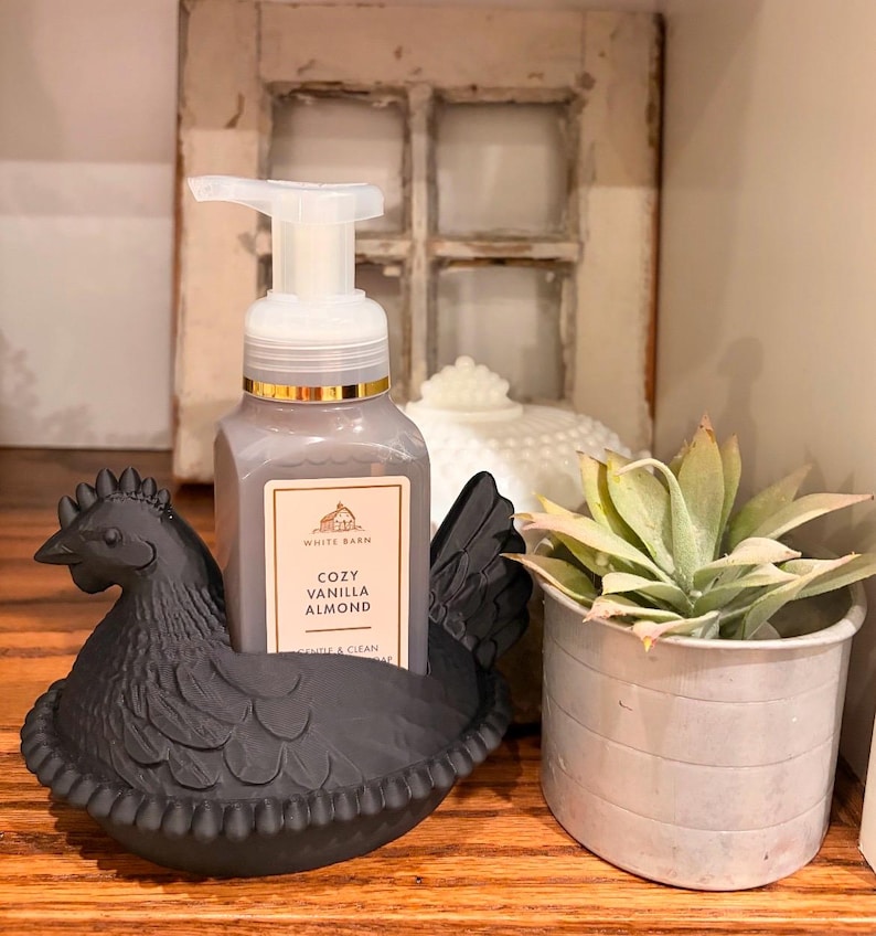 May include: A black hen-shaped soap dispenser holder cradles a bottle of Cozy Vanilla Almond soap. A small potted succulent and a decorative white object are also visible. The background includes a distressed window frame.